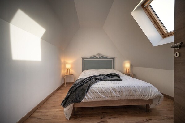 Un dormitorio con techo inclinado, una cama con colcha blanca y gris con lunares, dos lámparas de noche y un tragaluz.