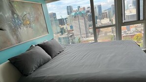 Ein Bett mit grauer Bettdecke und zwei Kissen, ein Gemälde an der Wand und ein Stadtblick durch große Fenster.
