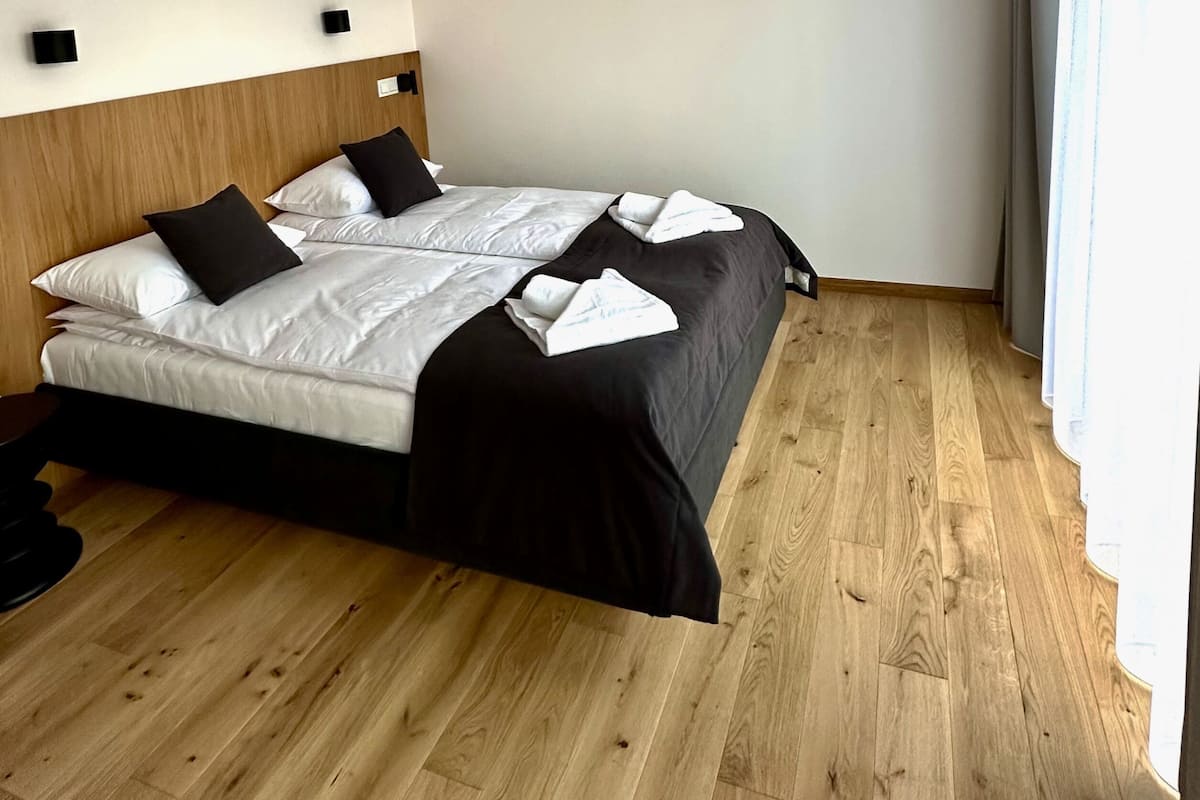 A modern hotel room with a wooden floor, a bed with white and black bedding, and a simple headboard.
