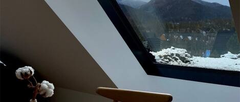 A room with a skylight, a vase with cotton, a plate with a croissant, and a view of snow-covered mountains.