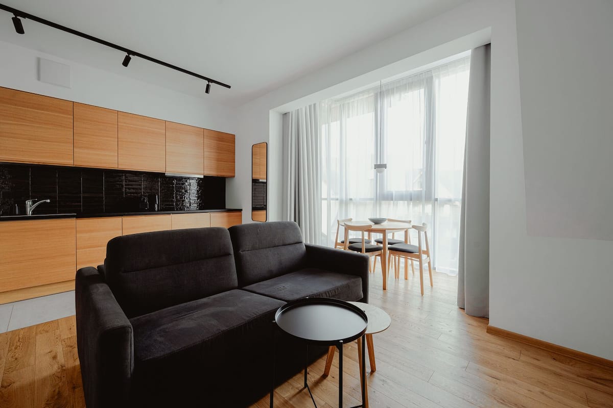 A modern living space with a sofa, a small round table, a dining area with chairs, and a kitchen with wooden cabinets and black countertops.
