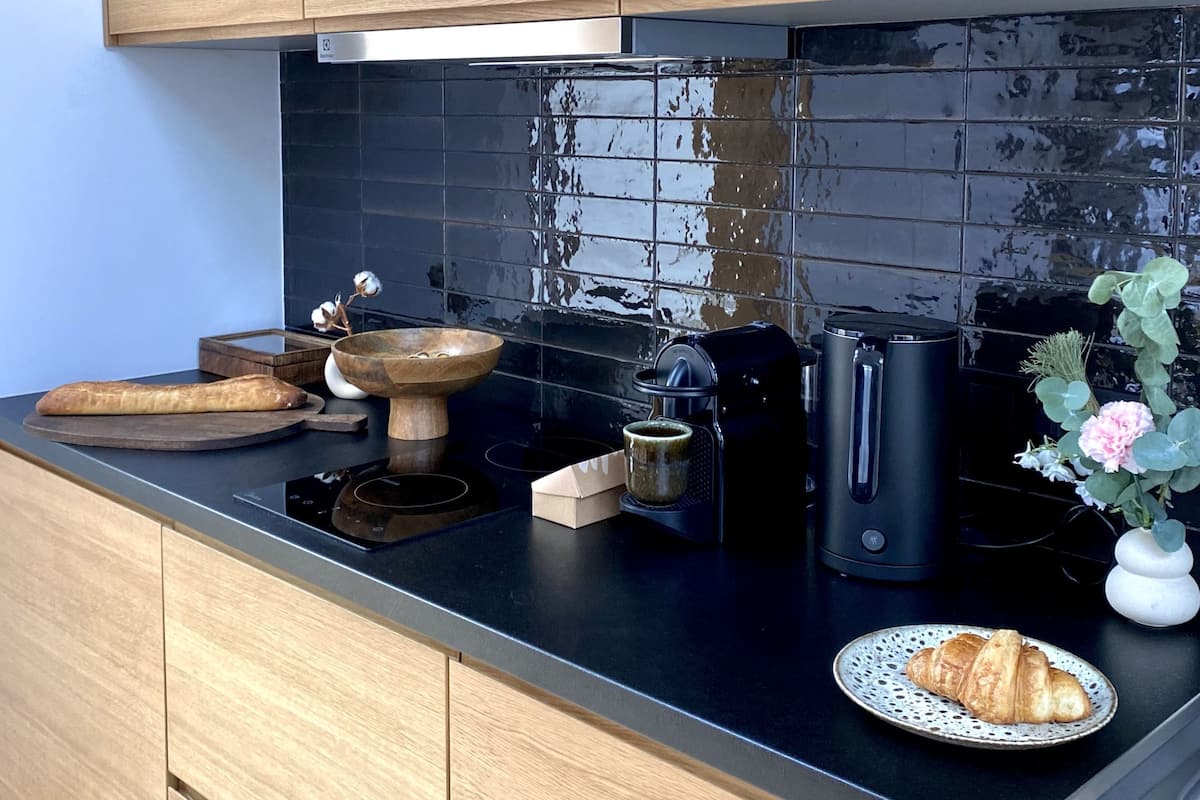 A modern kitchen with wooden cabinets, a black tiled backsplash, a coffee maker, a kettle, and a plate with croissants.