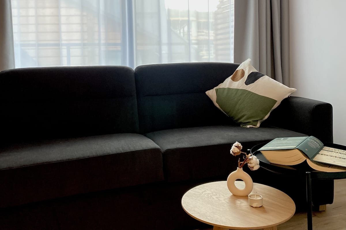 A modern living room with a dark sofa, a round wooden coffee table, and a window with sheer curtains.