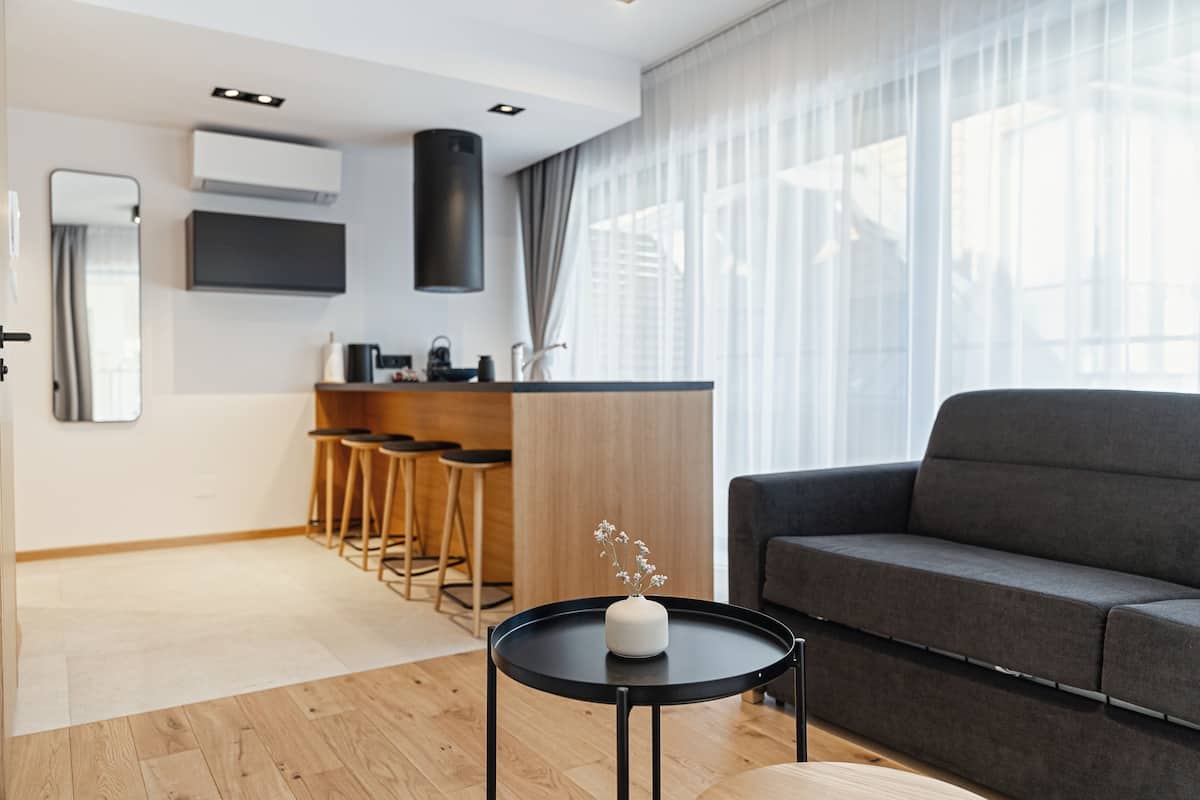 A modern living room with a grey sofa, a small black table with a vase, and a kitchen area with stools.