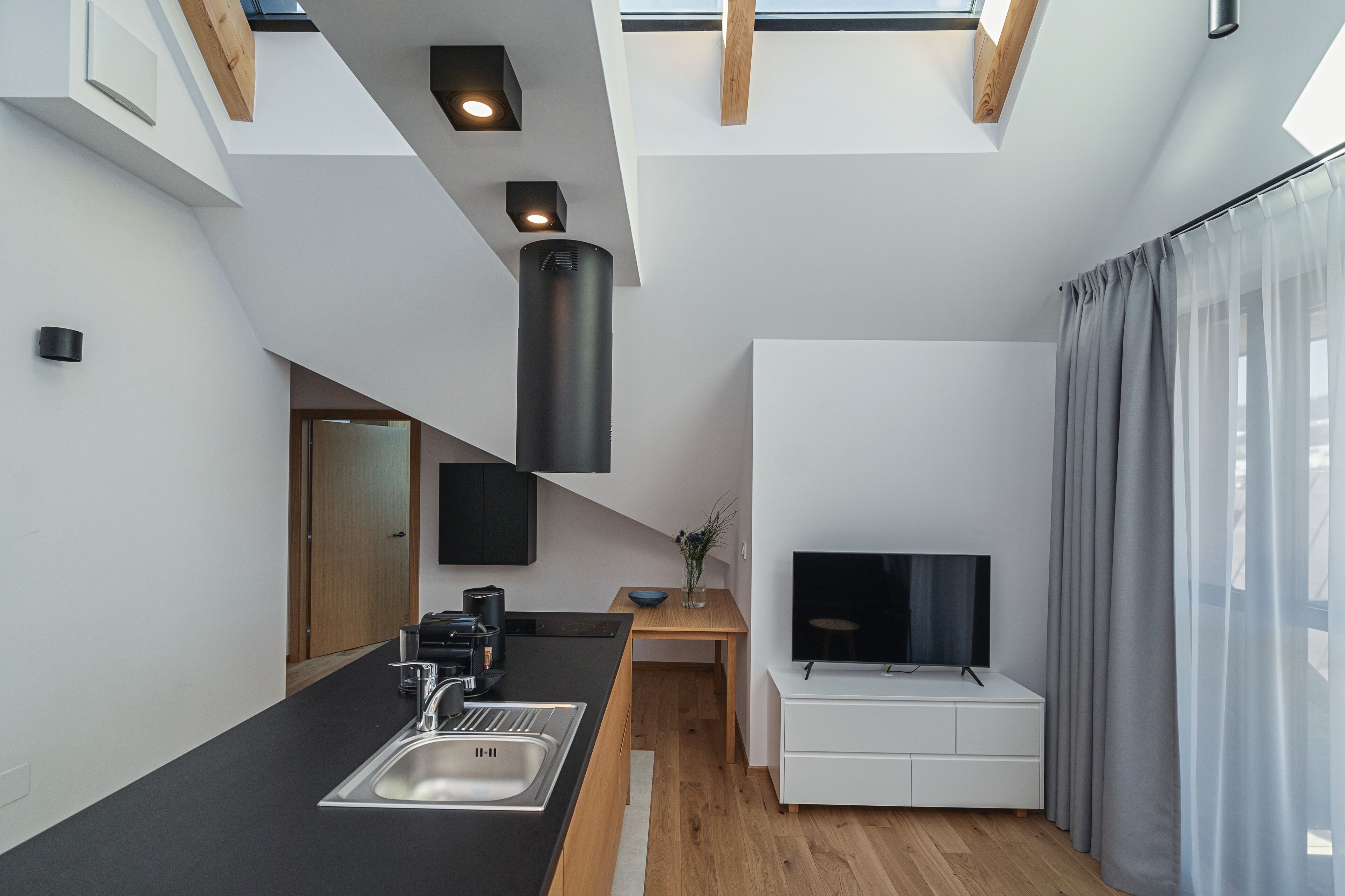 Una cocina moderna con encimera negra, pileta de acero inoxidable y estufa integrada. Hay una mesa de comedor de madera con un florero y un televisor de pantalla plana sobre un pedestal blanco.