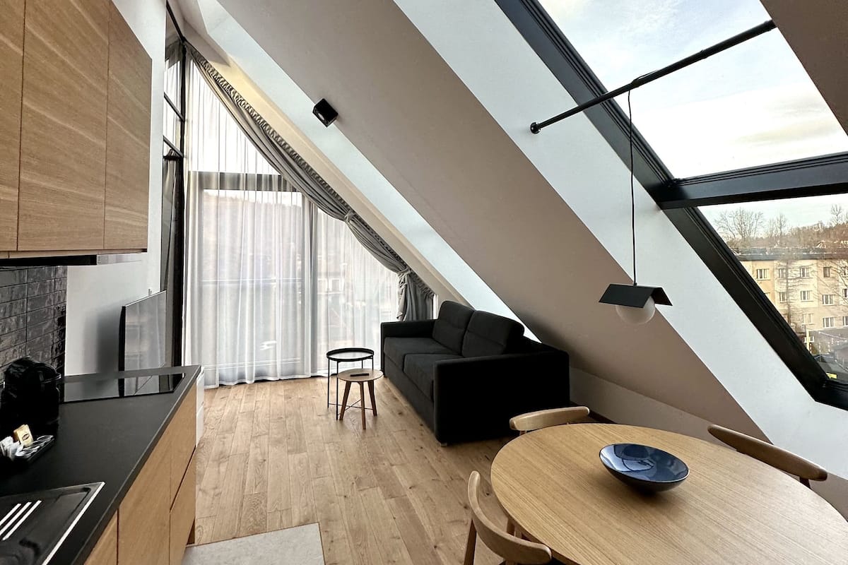 A modern loft apartment with a skylight, wooden flooring, a kitchen with a black countertop, a dining area with a round table and chairs, and a black sofa.