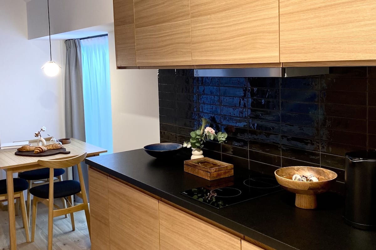 A modern kitchen with wooden cabinets, a black countertop, and a built-in stove. There is a dining area with a table and chairs, and a pendant light above.
