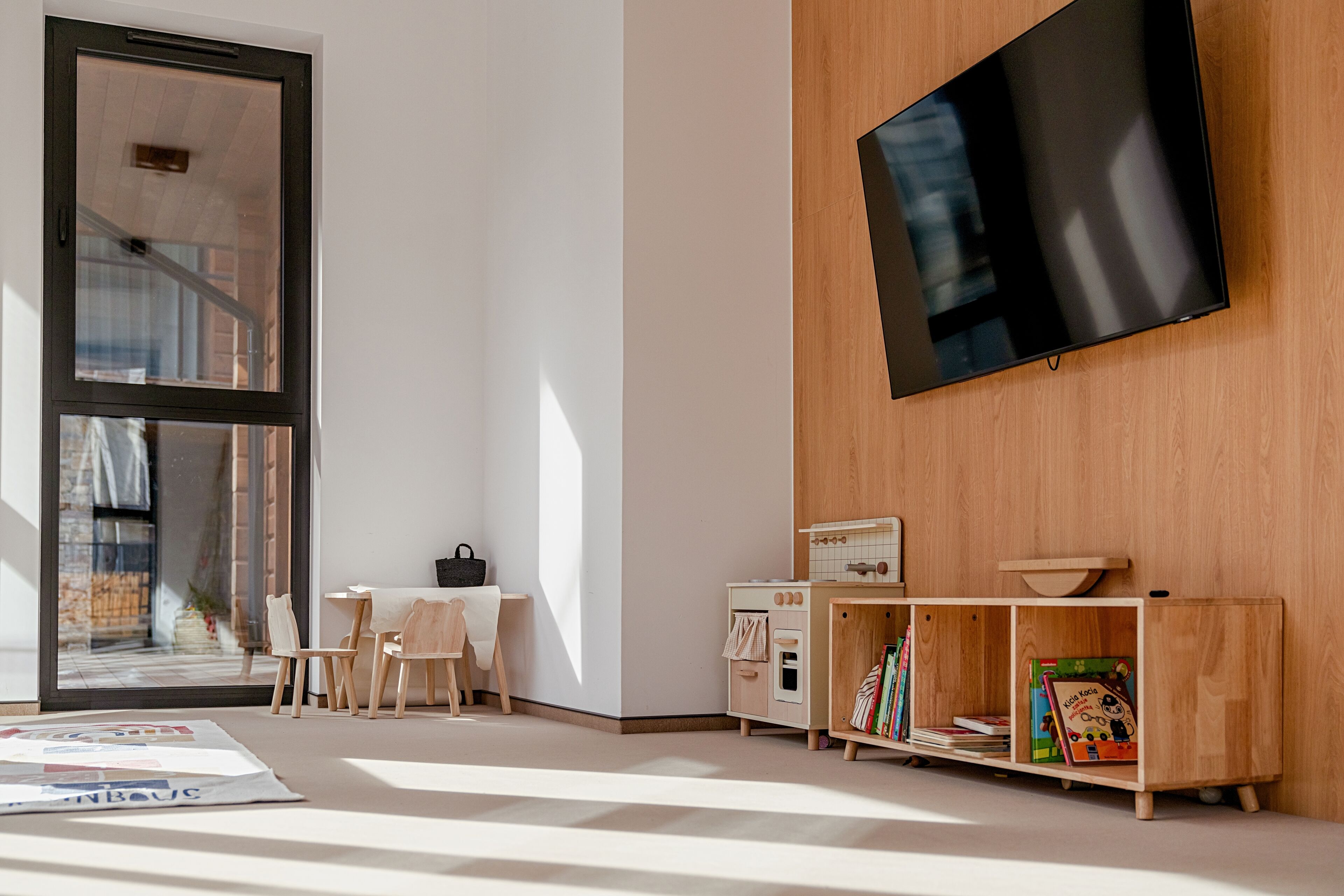 A modern living room with a large window, a flat-screen TV mounted on the wall, a wooden cabinet with books, and a small table with chairs.