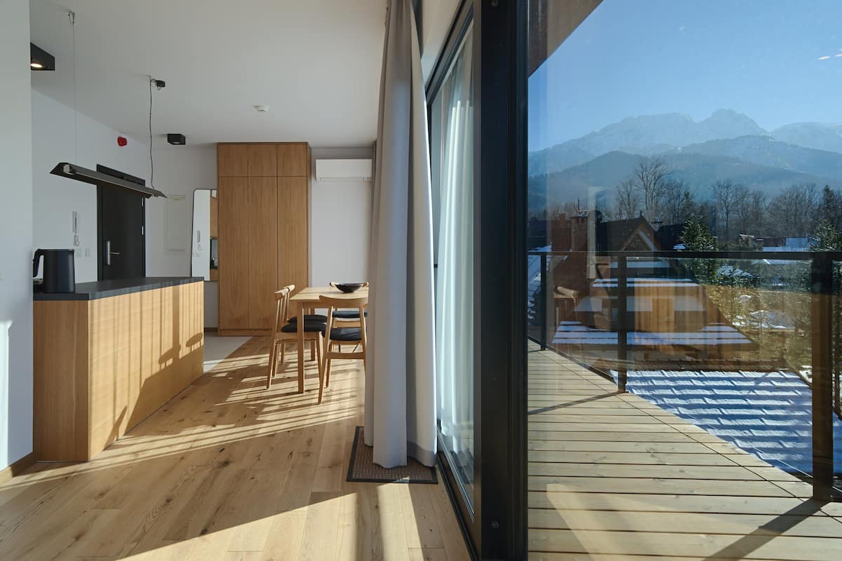 A modern kitchen with wooden cabinets and a dining area with a table and chairs. A large window offers a view of mountains.