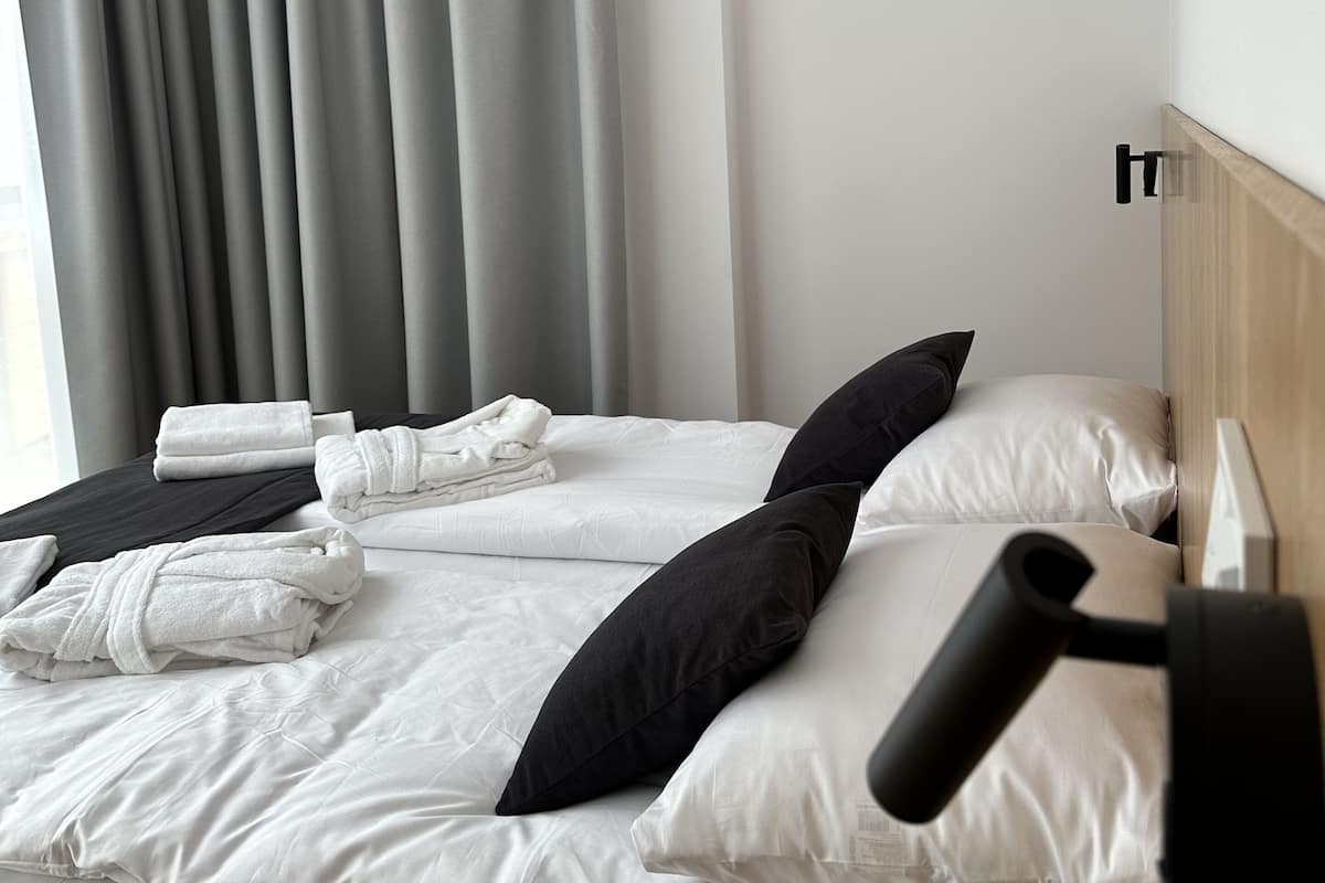 A hotel room with a neatly made bed, a set of towels, a black pillow, and a black lamp on the nightstand.