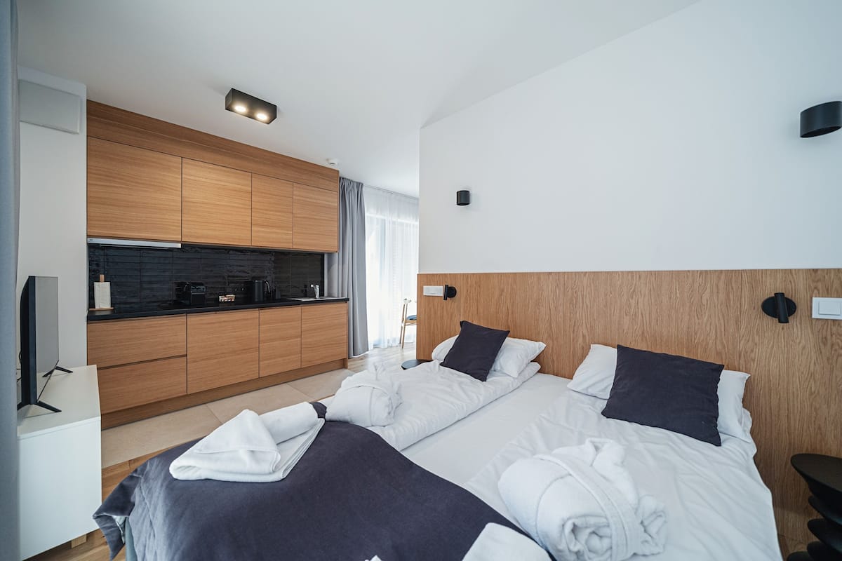 A modern hotel room with a large bed, a wooden headboard, a built-in kitchen with a black countertop, and a flat-screen TV.