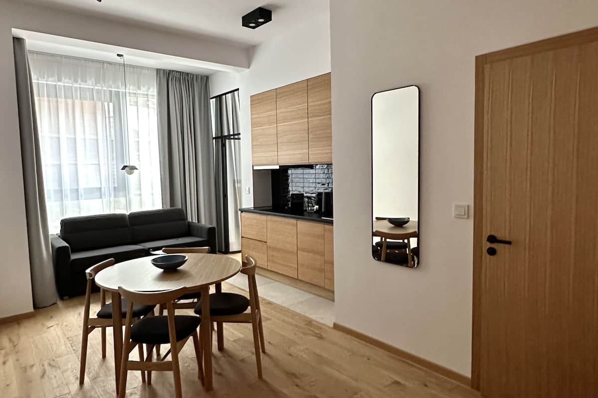 A modern living room with a dining table and chairs, a sofa, a kitchen area with wooden cabinets, and a large mirror on the wall.