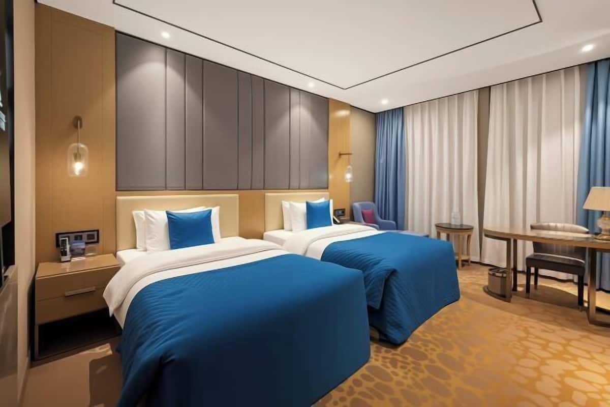 A hotel room with two beds, a desk, a chair, a small table, a lamp, and a curtain.