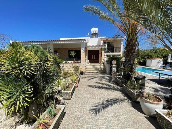A two-story house with a tiled patio leading to a front door, surrounded by various potted plants and a swimming pool.