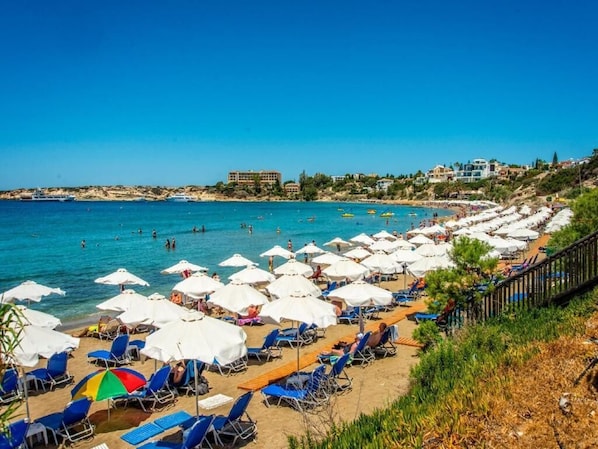 A beach with rows of sun loungers and umbrellas, a clear blue sea, and buildings in the background.