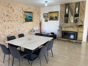 A dining area with a marble table and chairs, a brick wall with artwork, and a stone fireplace with a TV.