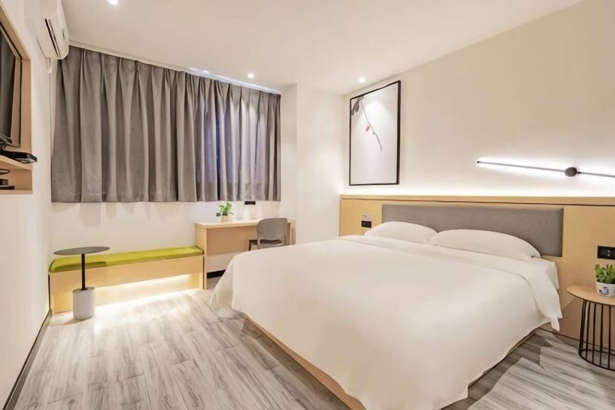 A modern hotel room with a large bed, a desk, a chair, a small table, a plant, and a large window with curtains.