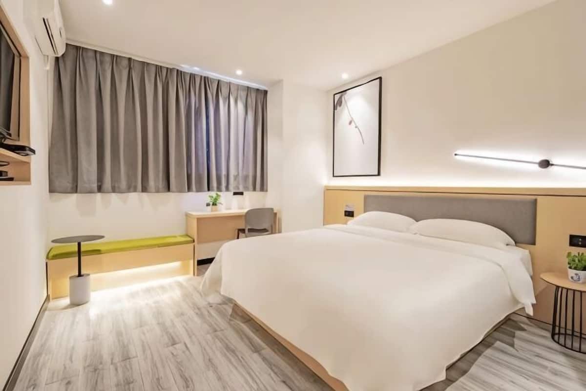 A modern hotel room with a large bed, a desk, a chair, a small table, a plant, and a large window with curtains.