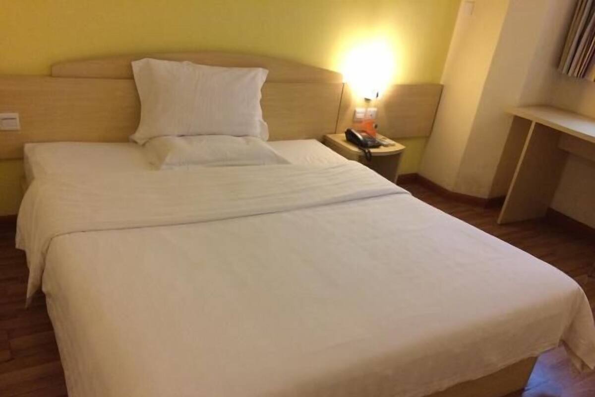 A bed with white linens, a wooden headboard, a bedside table with a lamp, and a small shelf.