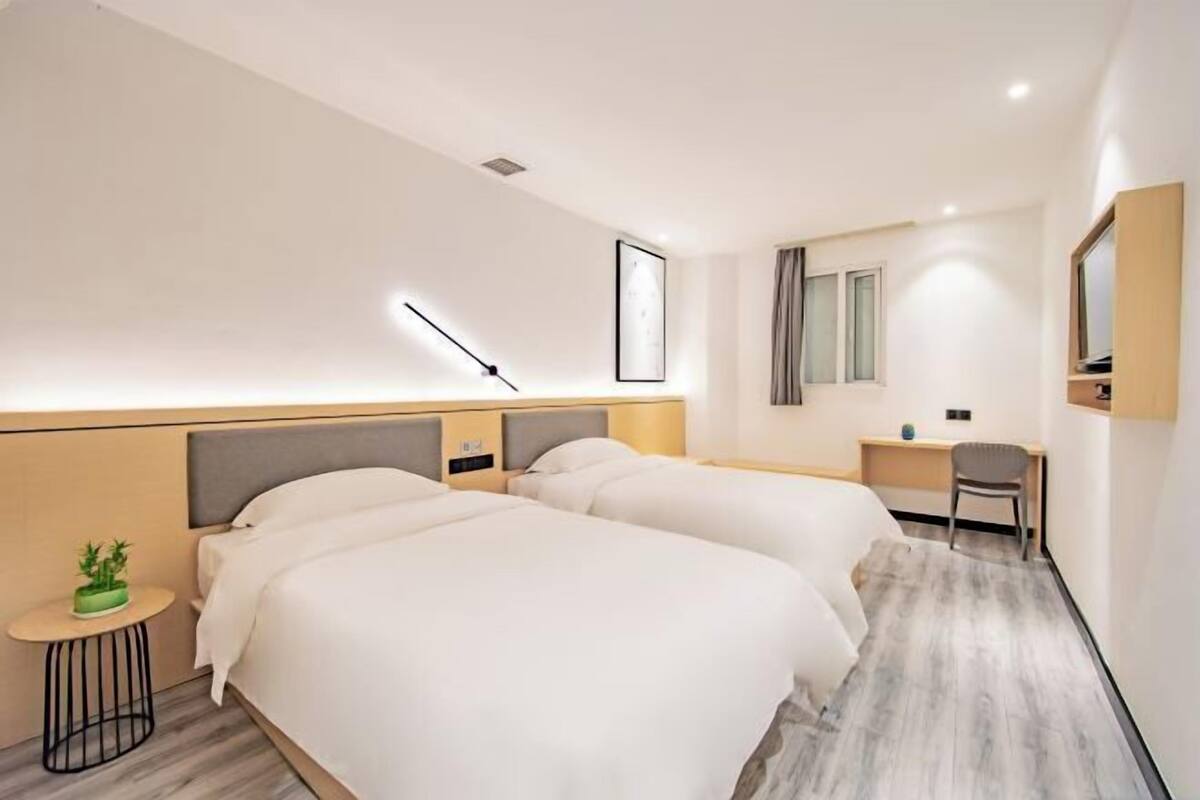 A hotel room with two beds, a desk, a chair, a small table with a plant, and a mirror on the wall.