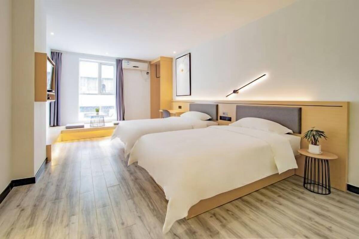 A hotel room with two beds, a small round table, a potted plant, and a window with curtains.