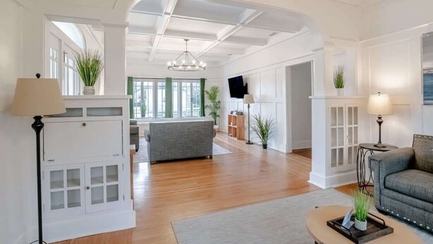 Un salón amplio con techo alto, piso de madera y un sofá gris. Hay dos lámparas, un televisor y una planta en el alféizar.