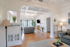Ein geräumiges Wohnzimmer mit hoher Decke, Holzboden und einem grauen Sofa. Zwei Lampen, ein Fernseher und eine Pflanze stehen auf der Fensterbank.