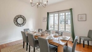 Comedor con una mesa de madera dispuesta para seis comensales, rodeada de sillas. Un candelabro cuelga del techo y hay dos ventanas con cortinas.