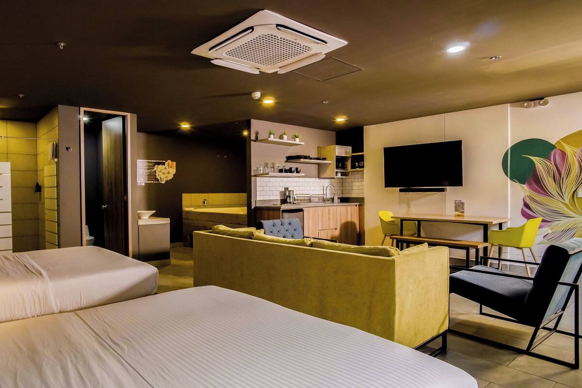 Habitació d'hotel moderna amb dos llits, una cuineta, una zona de menjador i un televisor.