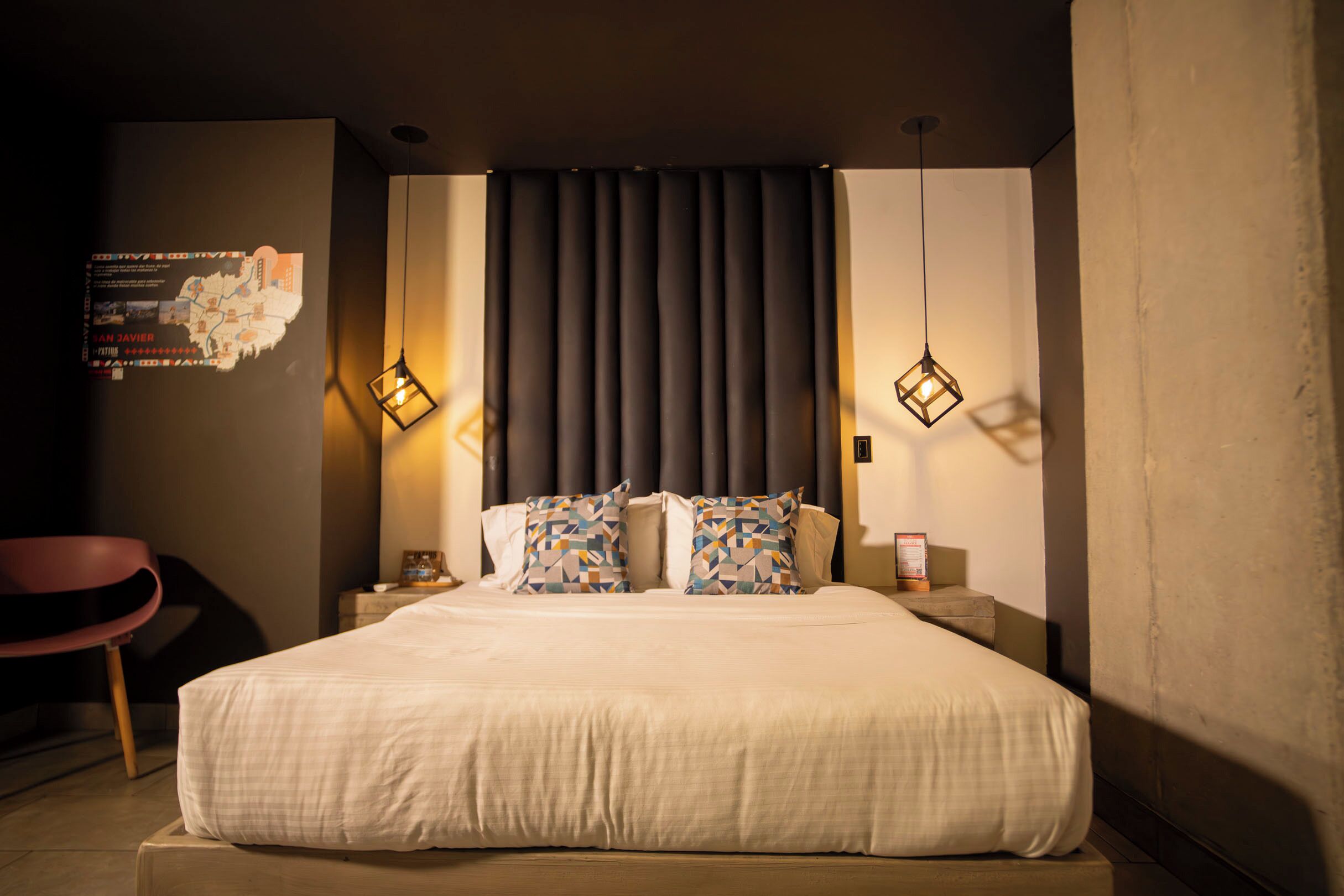 Una habitación de hotel moderna con una cama grande, almohadas con estampado geométrico, un cabecero oscuro y lámparas colgantes.