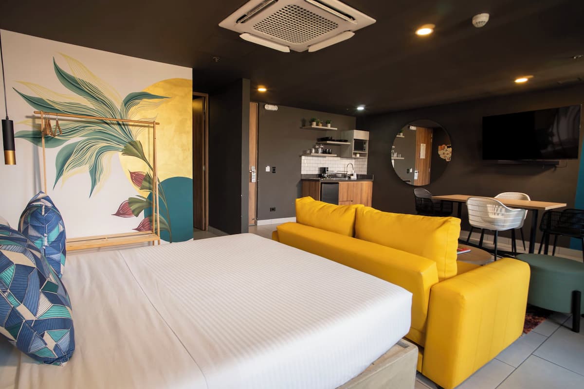 Habitació d'hotel moderna amb llit, sofà groc, zona de menjador i cuina al fons.