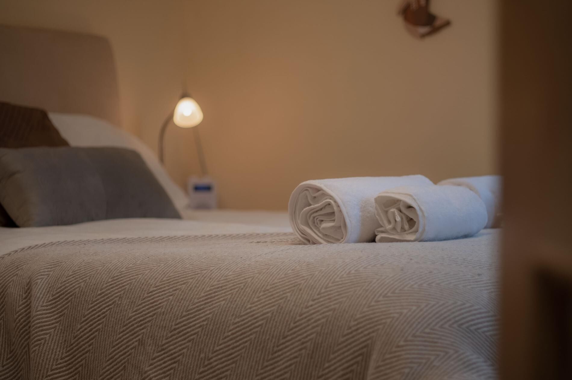 Ein ordentlich bezogenes Bett mit einem aufgerollten weißen Handtuch, einer Nachttischlampe und einem Kissen auf einer strukturierten Bettdecke.