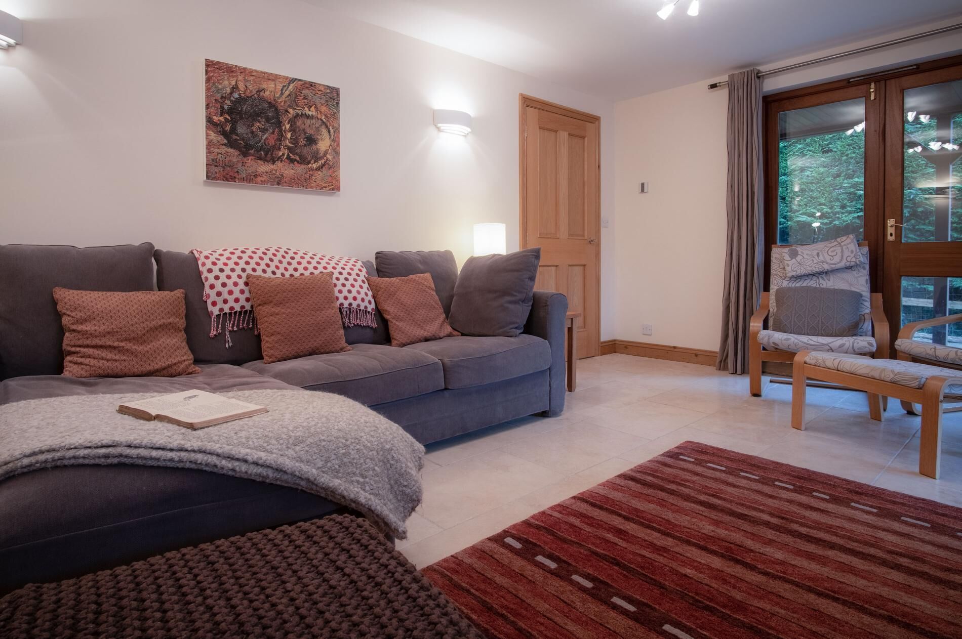 Un salon avec un canapé gris, des coussins à motifs, un plaid rouge et blanc à pois, un plaid tricoté, un tapis rouge à rayures et une chaise en bois avec un coussin.