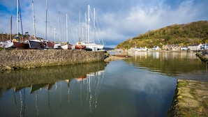 Un port de plaisance avec des bateaux amarrés le long d’une jetée en pierre, des eaux calmes qui reflètent les bateaux, et une colline avec des bâtiments en arrière-plan.
