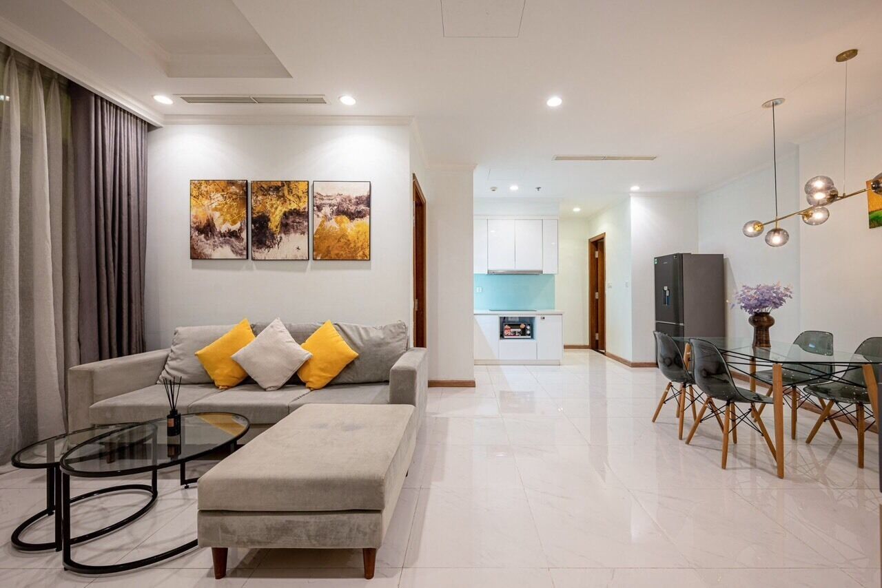 A modern living room with a sofa, ottoman, and glass coffee table. There is a dining area with a glass table and chairs. The room has a kitchen in the background.
