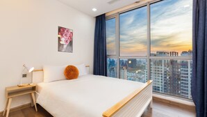 Una habitación de hotel moderna con una cama grande, una mesita de noche, una lámpara de escritorio, vistas a la ciudad a través de amplios ventanales y una obra de arte floral enmarcada en la pared.