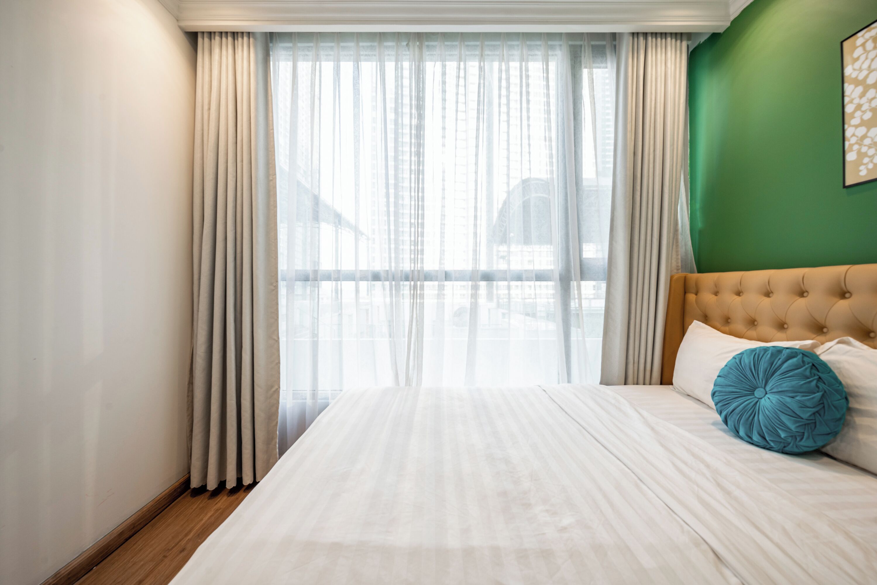 A hotel room with a large bed, beige curtains, a green accent wall, and a view through sheer curtains.