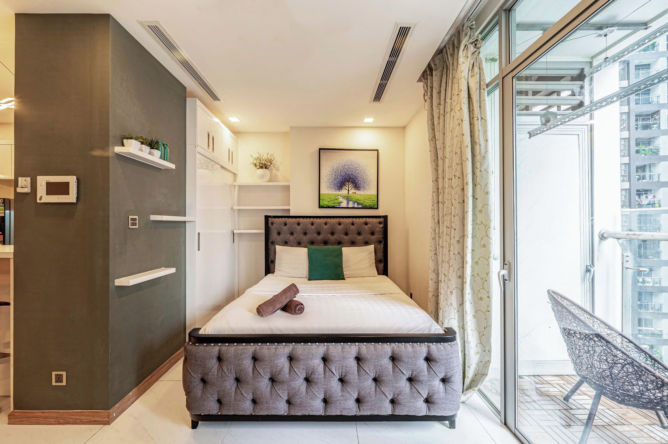 Un dormitorio con cama de cabecera capitonada, mesita de noche, armario empotrado, pequeña estantería con plantas, un cuadro de un árbol y vistas a un balcón con una silla.