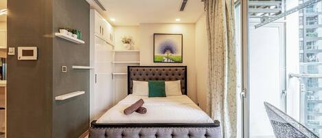 Un dormitorio con cama de cabecera capitonada, mesita de noche, armario empotrado, pequeña estantería con plantas, un cuadro de un árbol y vistas a un balcón con una silla.