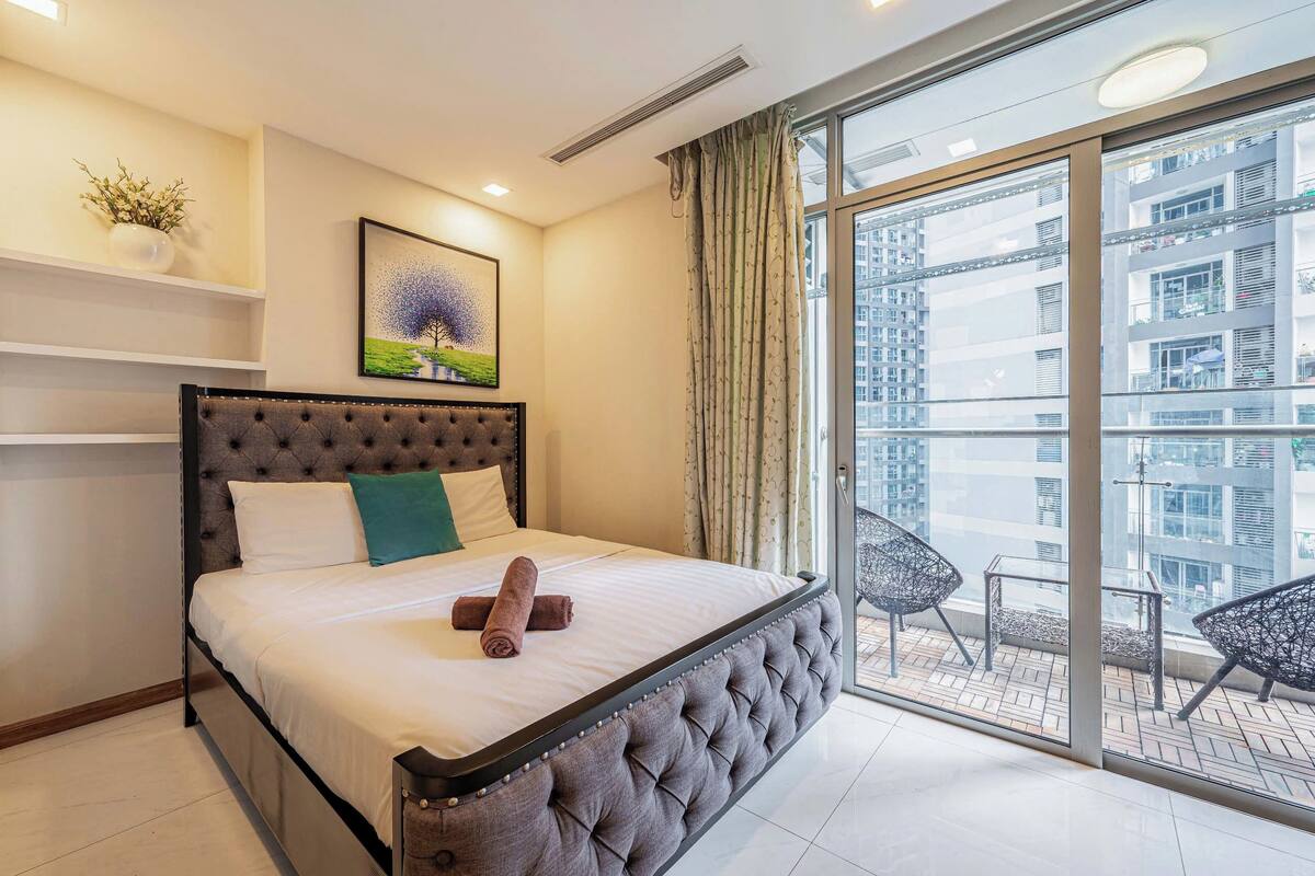 A bedroom with a large bed, a window with a view of buildings, a potted plant, and a framed picture on the wall.