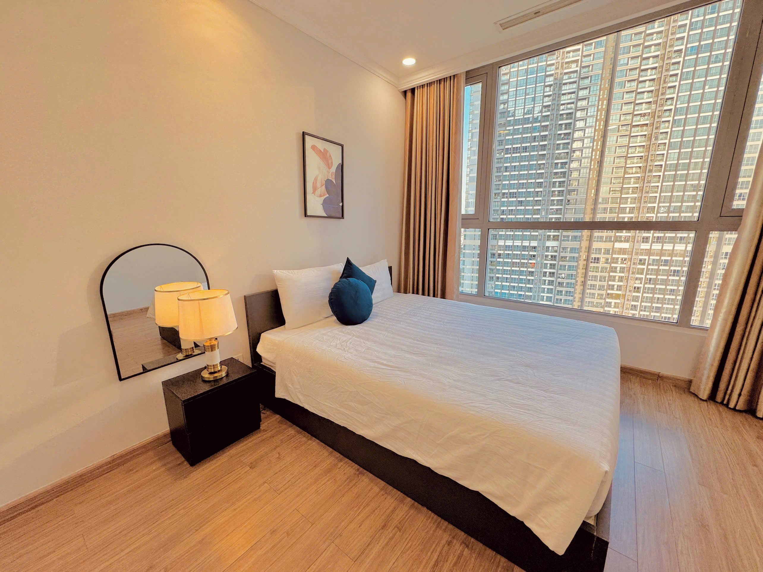 A hotel room with a large bed, a bedside lamp, a mirror, and a window with a city view.