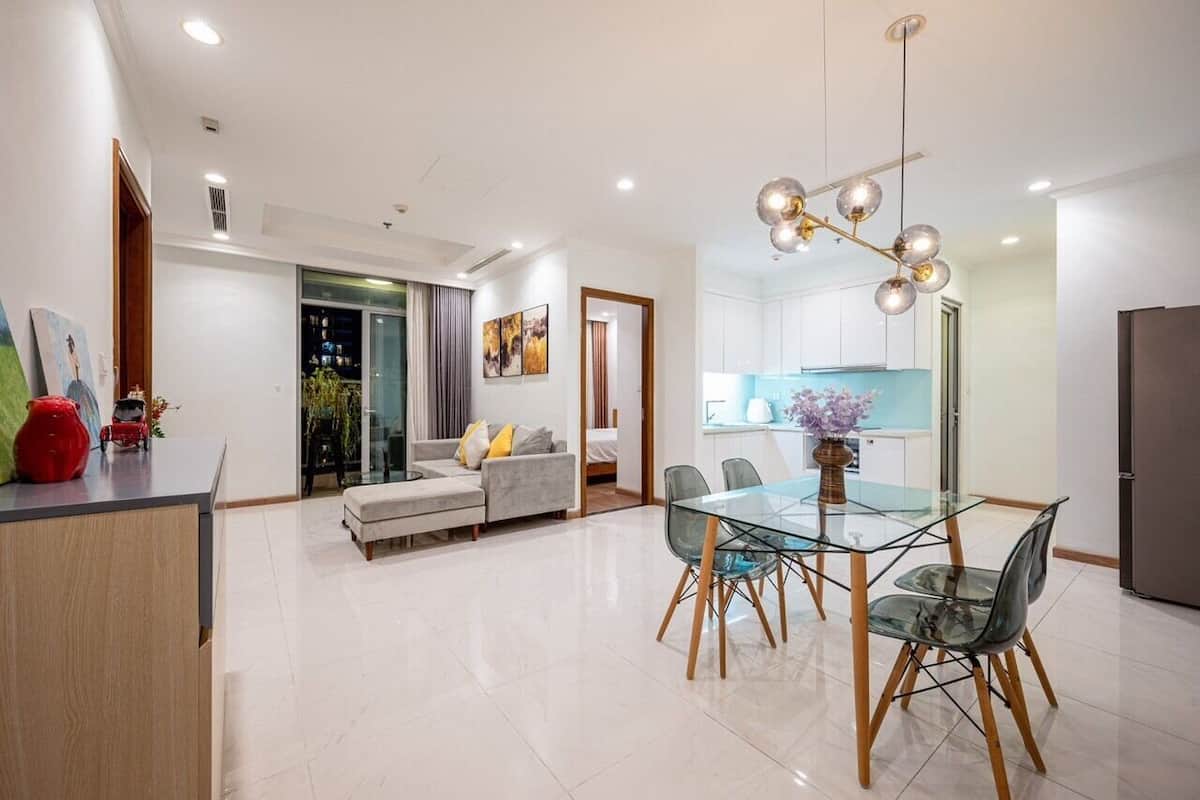 A modern living room with a glass dining table, grey sofa, and a kitchen in the background.