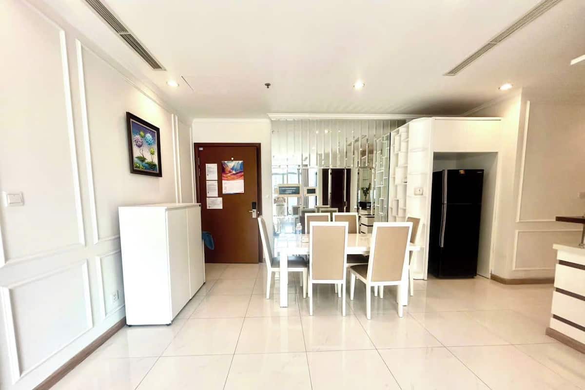 A spacious, well-lit dining area with a modern white table and chairs, a refrigerator, and a kitchen area visible in the background.