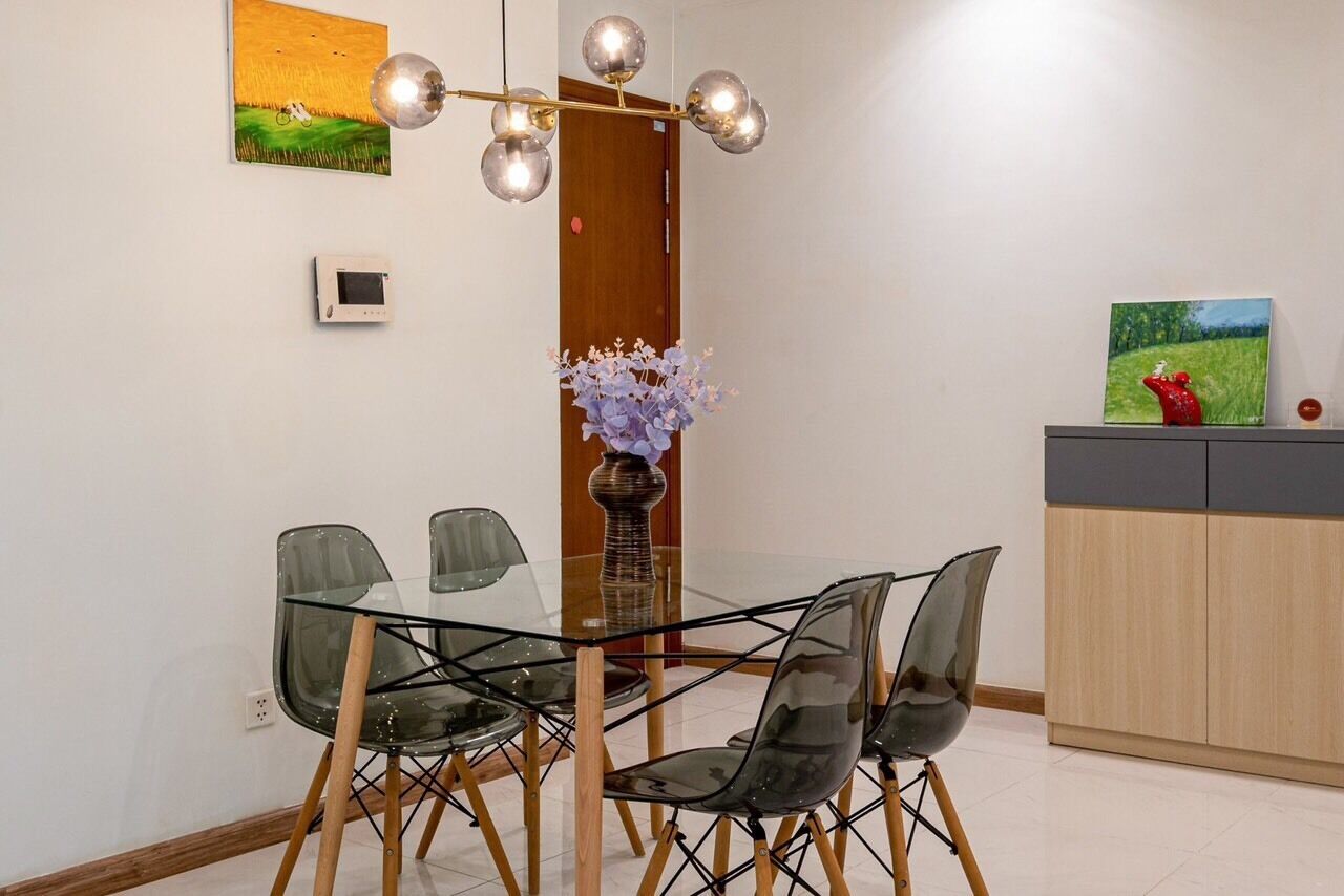 A dining area with a glass table and chairs, a vase with flowers, a wall-mounted thermostat, and a painting above the table.