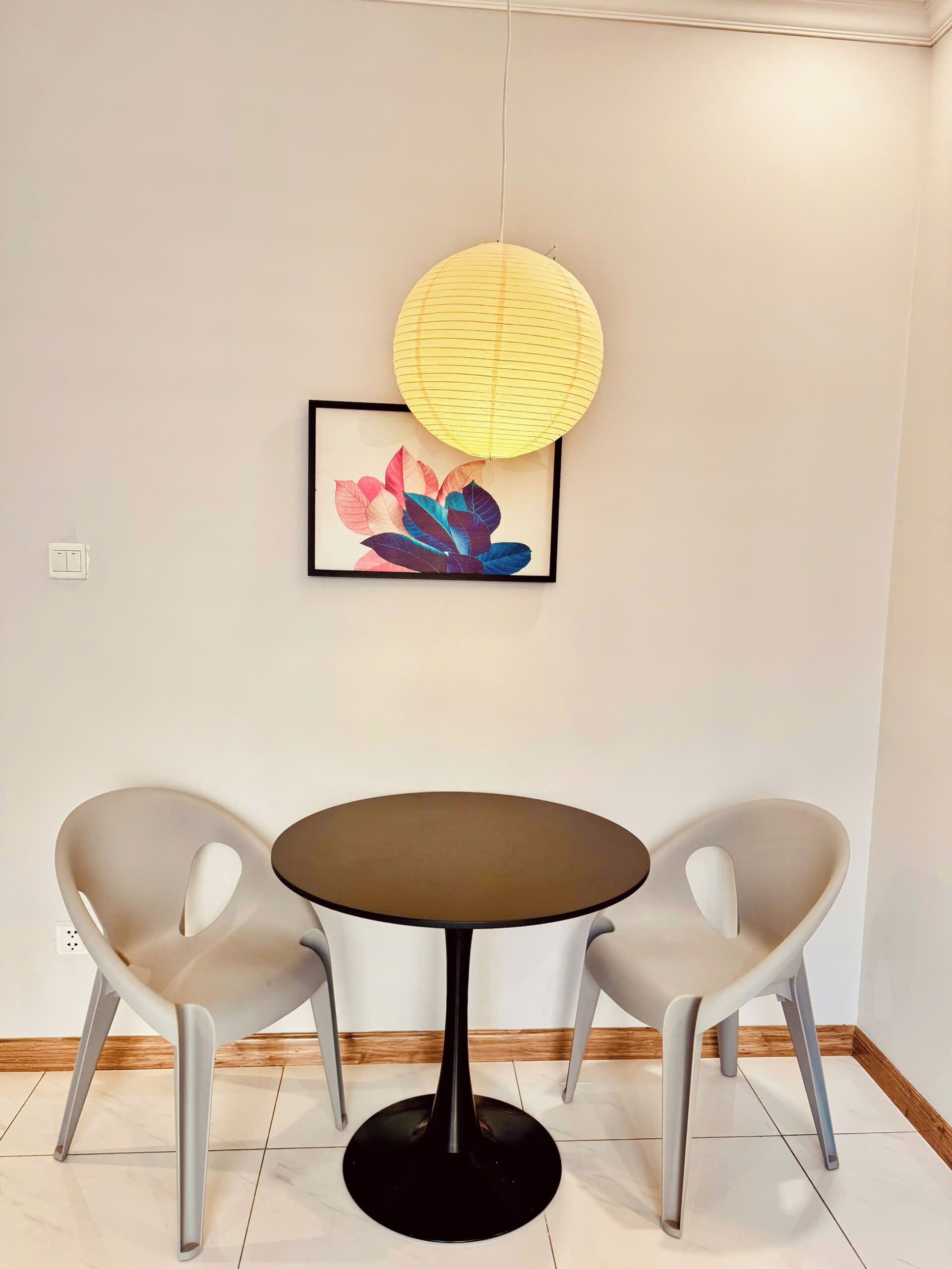 A room with a round table and two chairs, a framed artwork on the wall, and a hanging paper lantern.