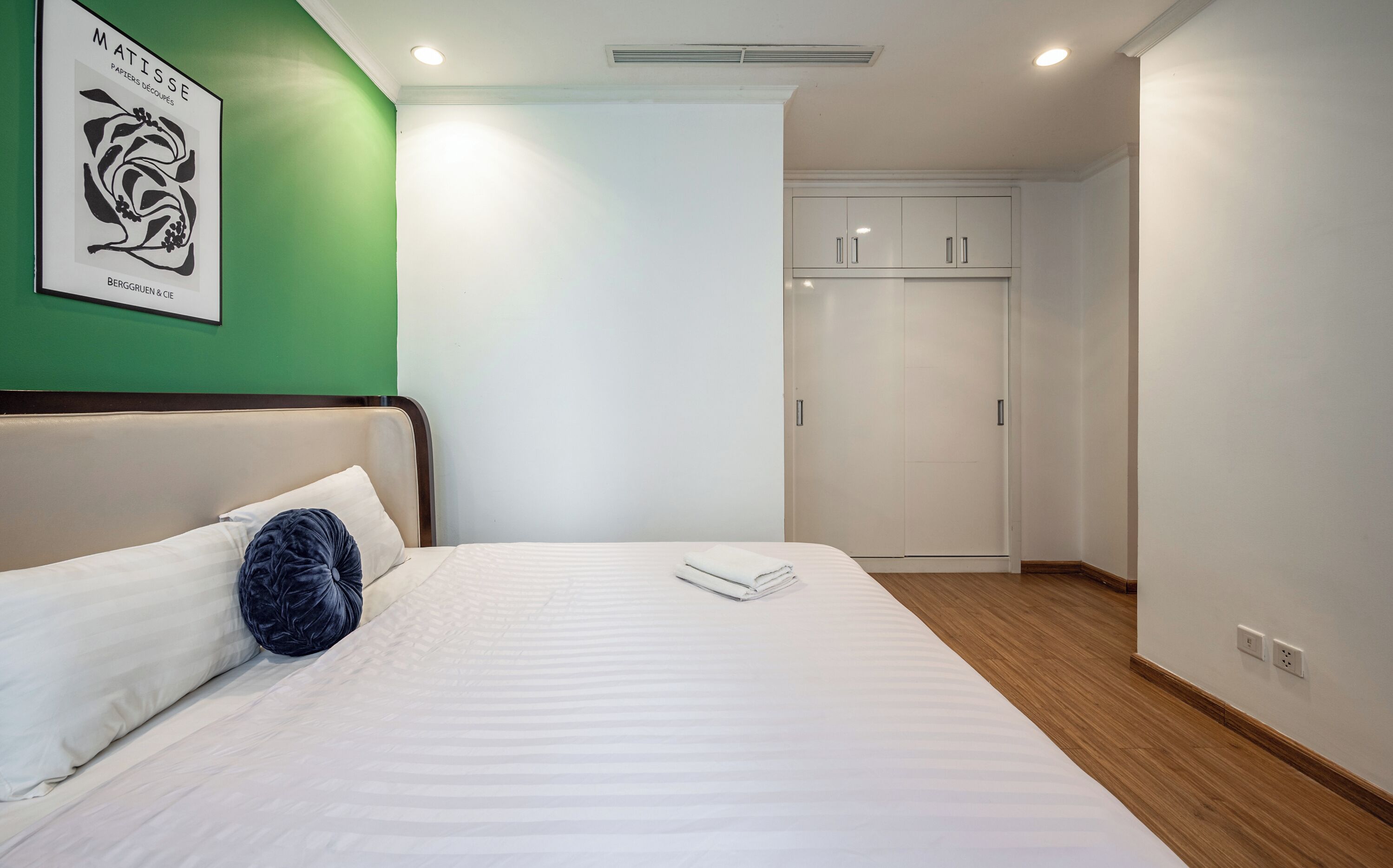 Una cama bien hecha con ropa blanca y una almohada decorativa azul, un armario blanco con espejo y una pared de acento verde.