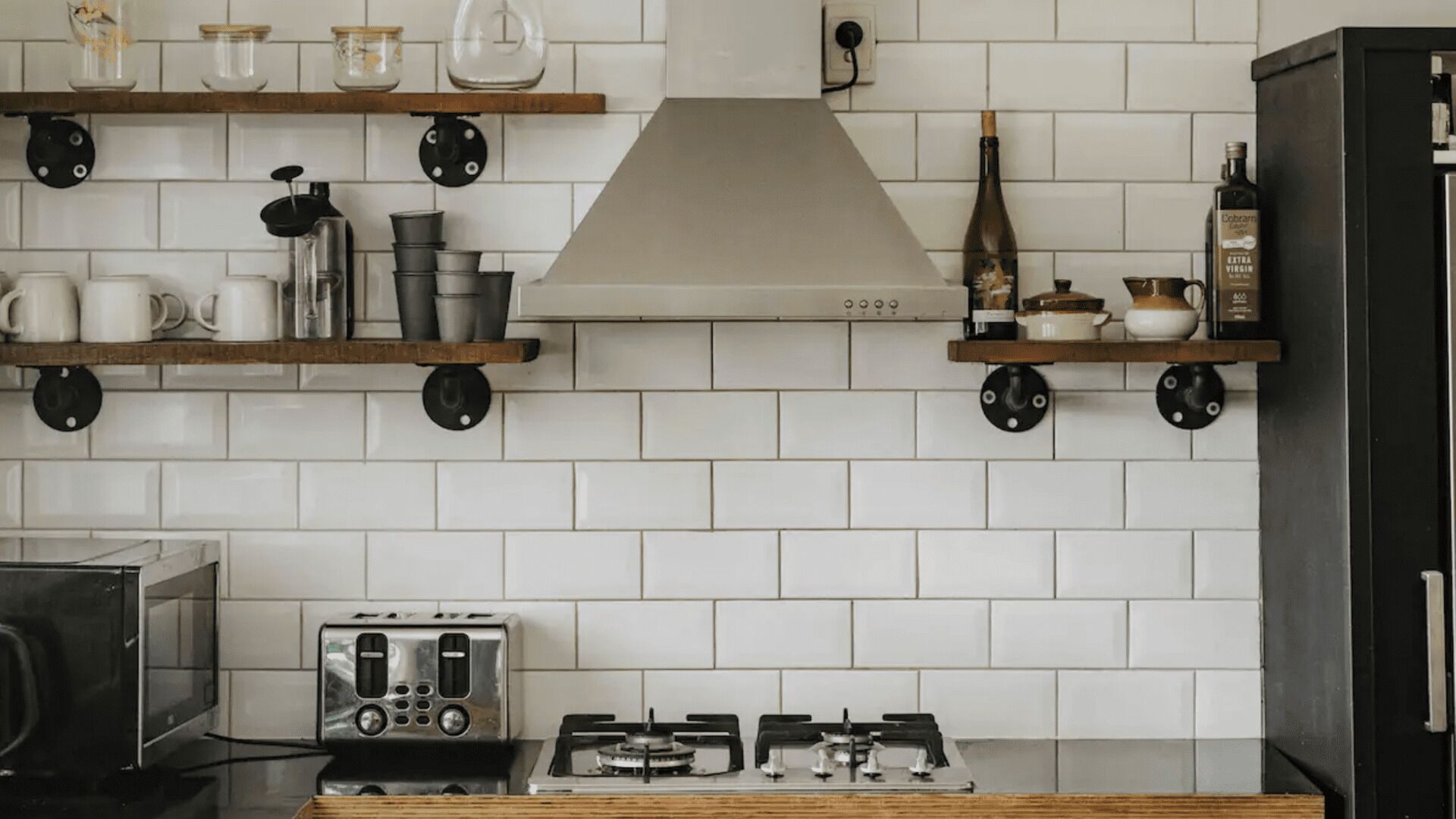 Een keuken met een witte betegelde achterwand, roestvrijstalen apparatuur, houten planken en een gasfornuis.