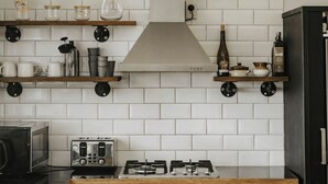 A kitchen with white tiled backsplash, stainless steel appliances, wooden shelves, and a gas stove.