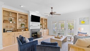 A spacious living room with a fireplace, blue sofas, a television, and a large window with a view of greenery. - 3 Summer Dunes - Renovated Ocean View Beach House! (Isle of Palms)