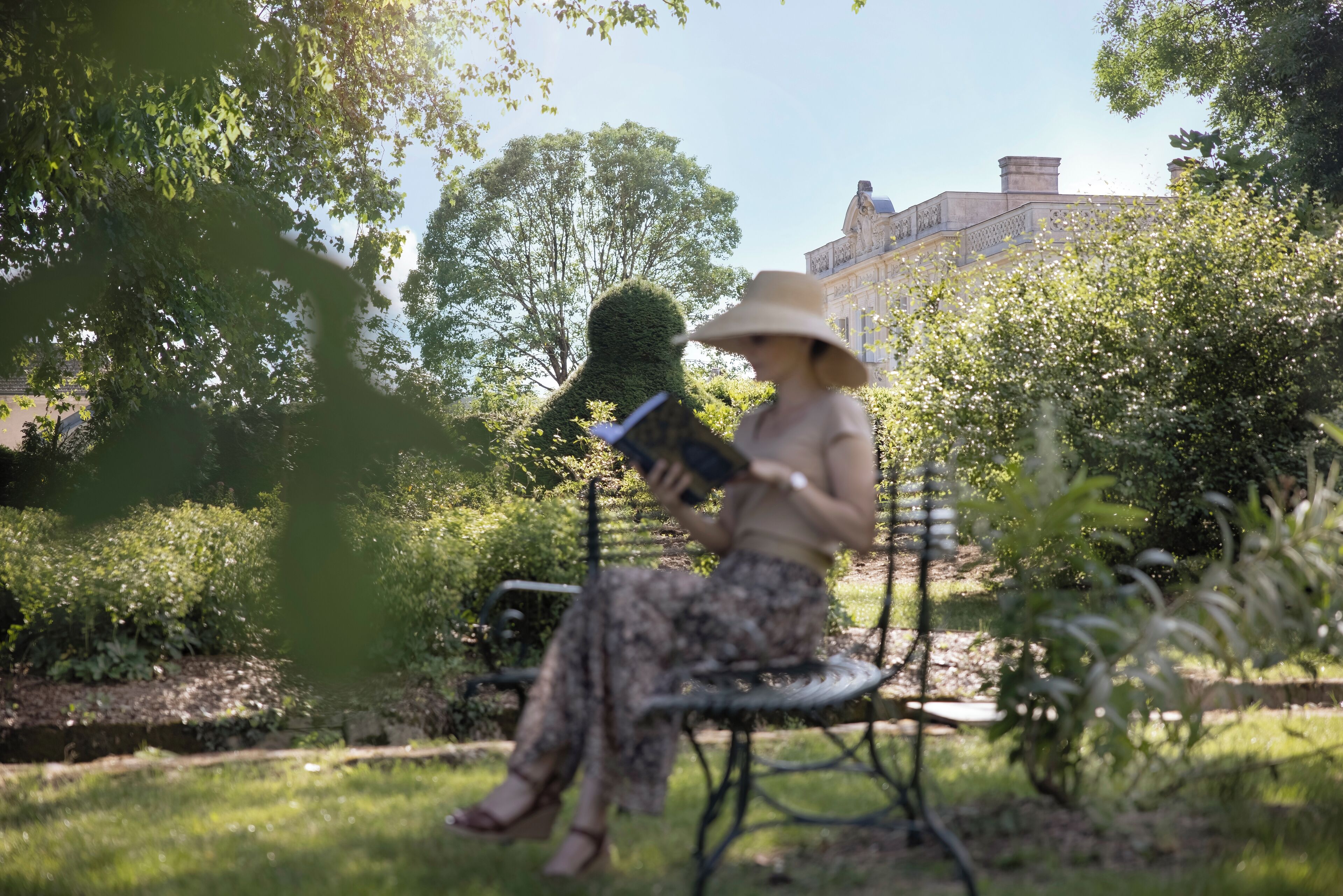 Une personne assise sur un banc dans un jardin, avec un bâtiment en arrière-plan.